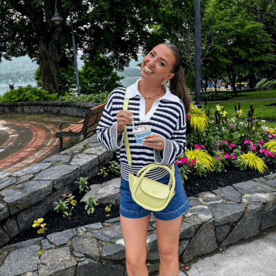 A smiling woman stands outdoors by a flower bed and stone wall near Lake George, holding a drink. She wears a striped sweater, denim shorts, and a light yellow purse. Trees and a park bench are visible in the background.